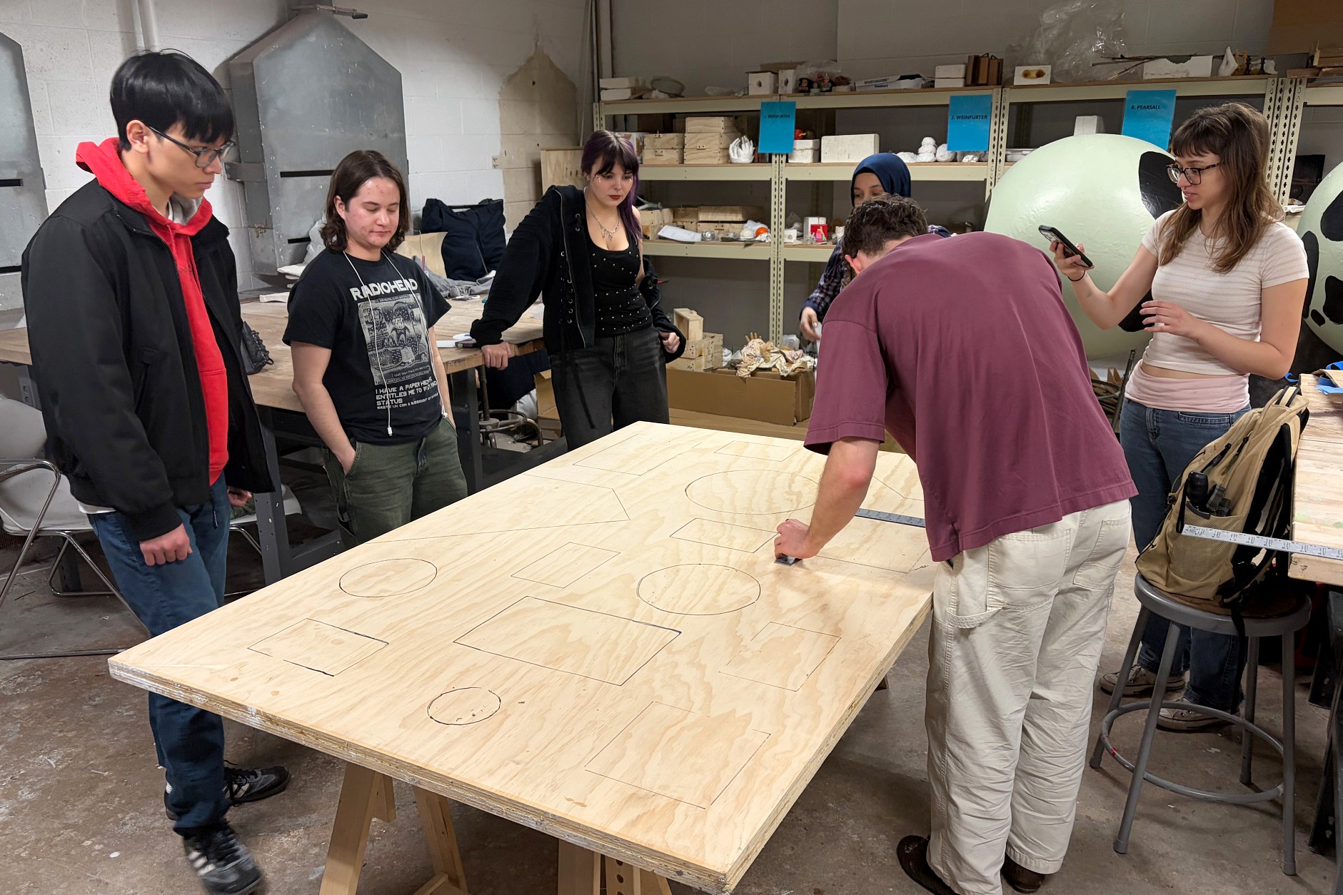 A group of six people in a workshop observe a man drawing shapes on a large plywood sheet. The setting is creative and collaborative.