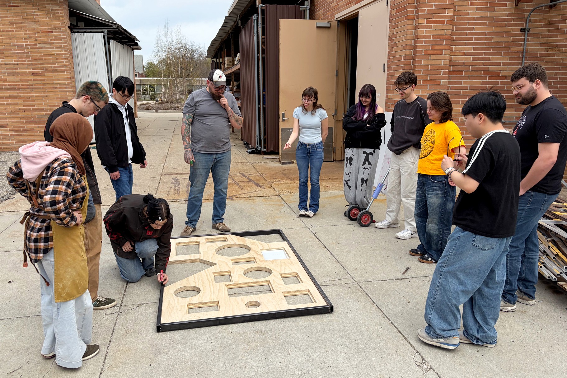 A group of students and an instructor observe a large wooden puzzle on the ground outside a workshop. The mood is curious and collaborative.