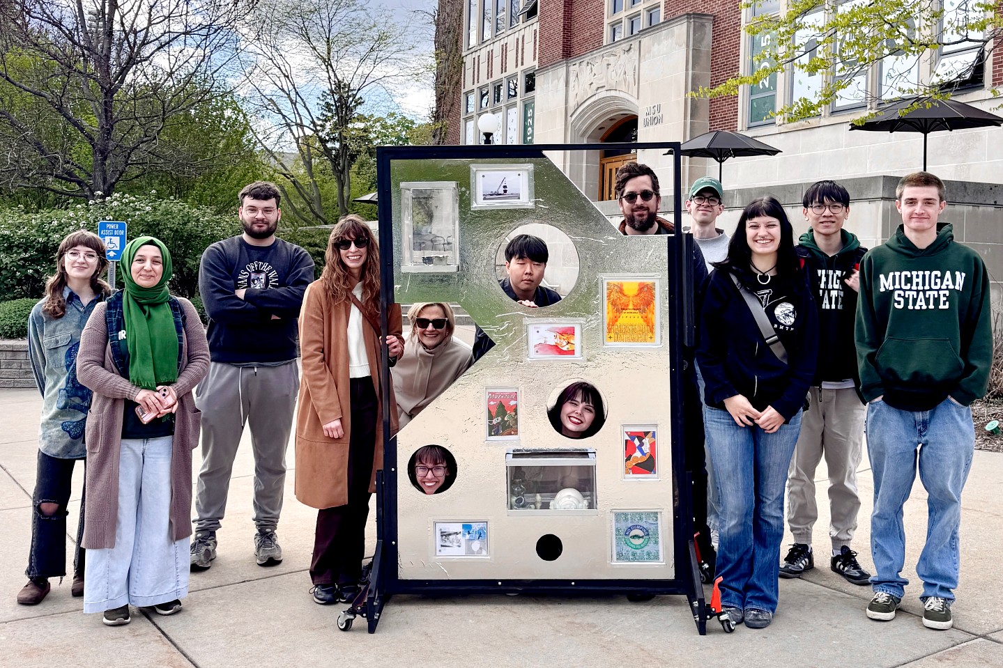 A group of students stand smiling outdoors, surrounding an art display featuring small artworks and circular cutouts for faces, against a university building backdrop.