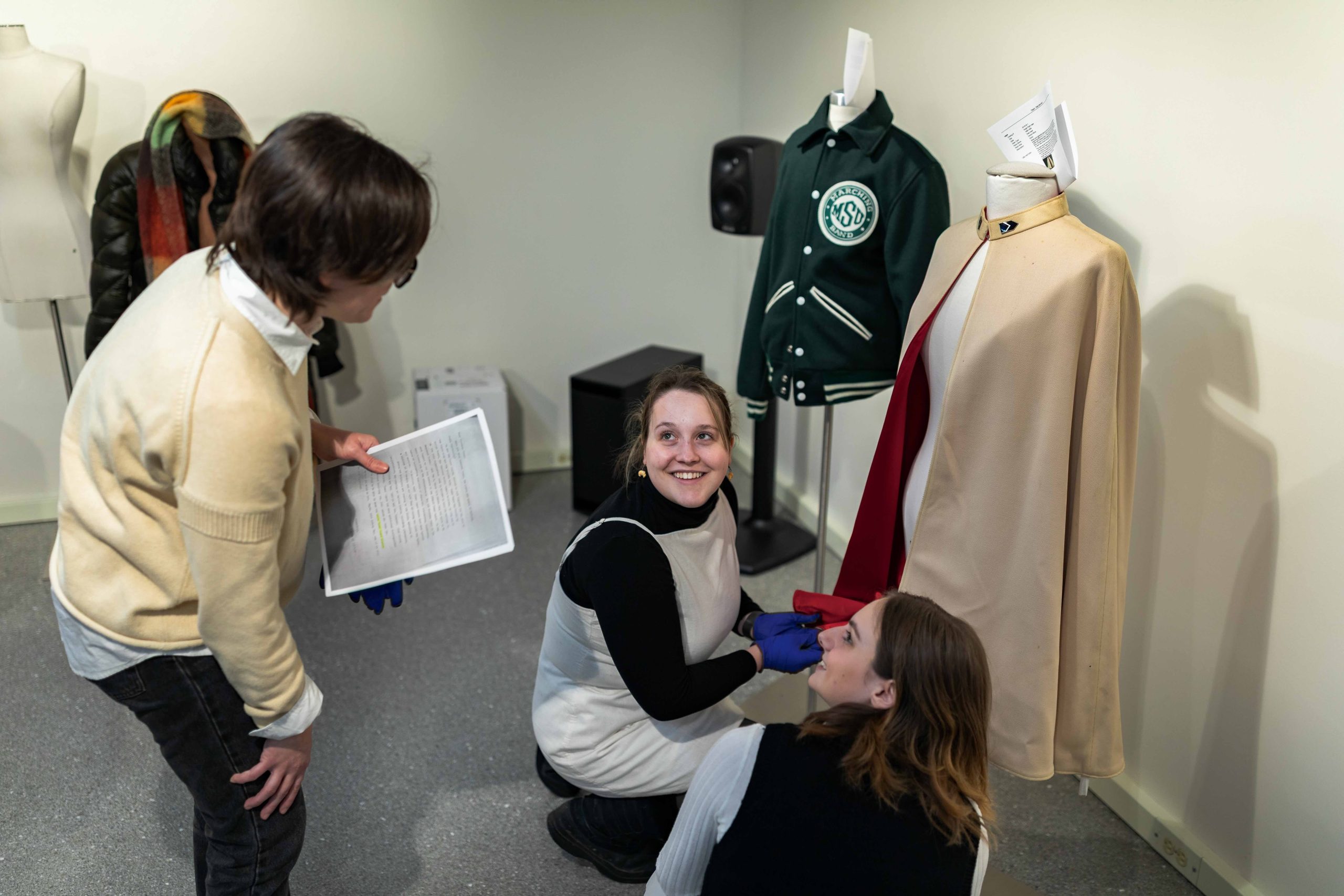 Two students crouch down in front of a dress form showcasing a cream jacket. Another student stands over them, engaging in a discussion.