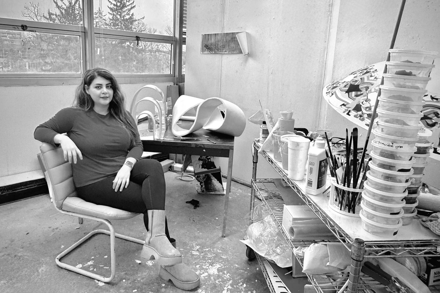A woman sits in an art studio with modern sculptures on a table, surrounded by art supplies. Large windows in the background.