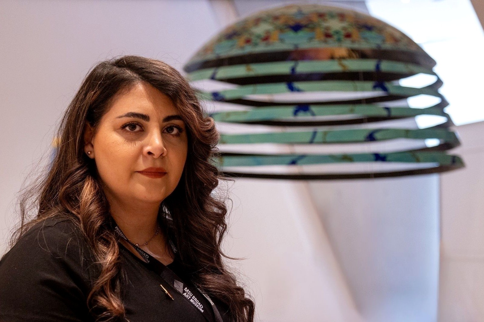 A woman with long brown hair stands indoors, looking at the camera with a composed expression. Behind her is a colorful, layered sculpture.