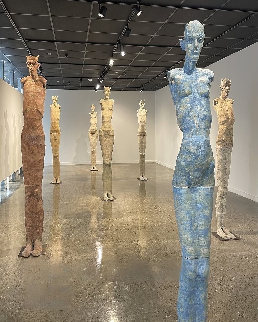A gallery featuring nine tall, elongated human-like sculptures in earthy tones. They stand on a polished floor under spotlights, creating a contemplative atmosphere.