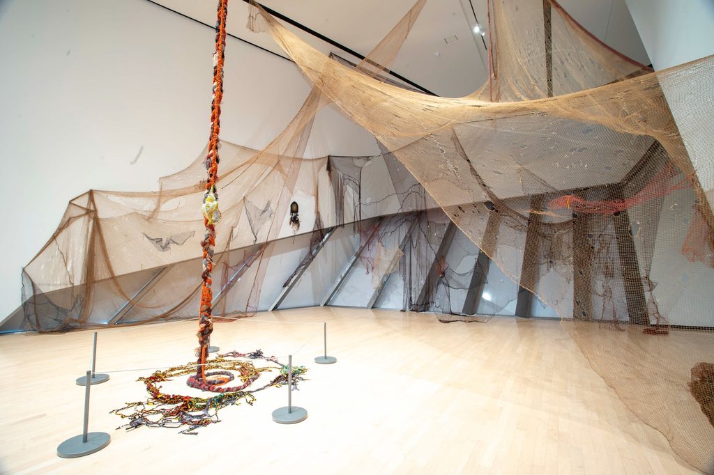 Installation art with large interconnected nets draping from the ceiling, a central rope wrapped in colorful fabric, creating a spiral pattern on the floor.