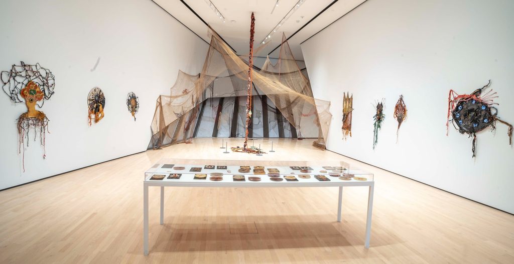 A gallery with white walls showcasing abstract installations. A table with art pieces in the foreground. Large netted structures and wall art evoke curiosity.