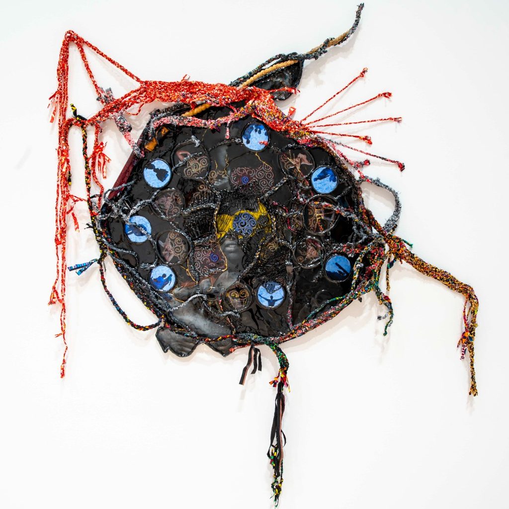 Abstract textile art featuring a circular centerpiece adorned with blue and brown patterned circles, surrounded by intertwined red and multicolored threads, creating a dynamic, chaotic feel.