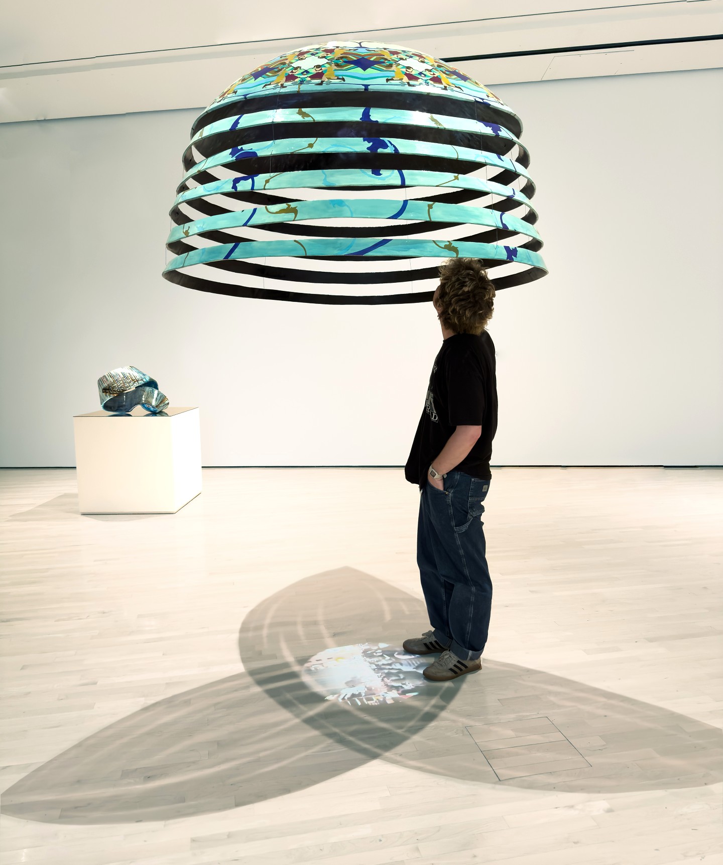 A person stands beneath a large, blue and black striped dome-shaped art installation, casting complex shadows on the gallery floor. A second artwork sits nearby on a pedestal.