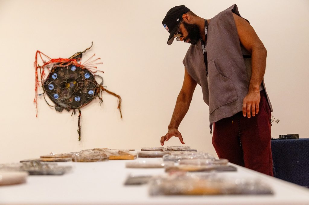 A man wearing a cap and a sleeveless shirt examines art pieces on a table in a gallery. A colorful, abstract wall sculpture is visible in the background.