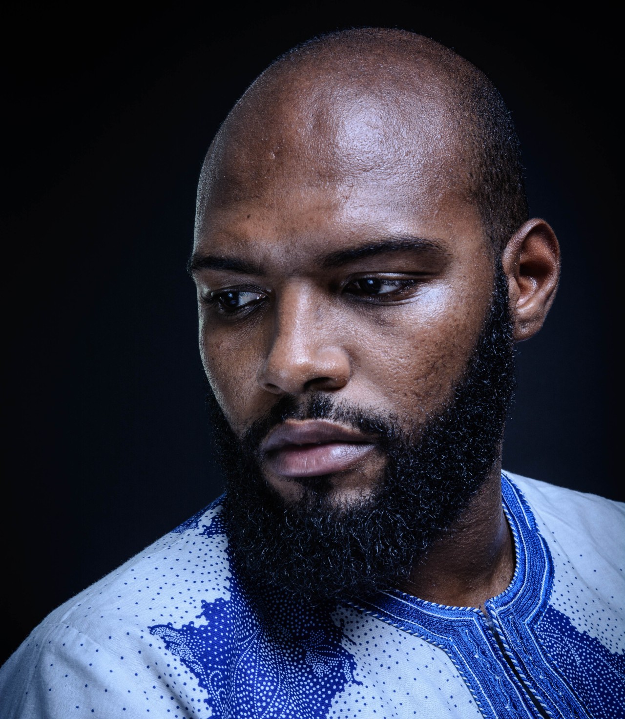 Portrait of a man with a thoughtful expression, wearing a blue embroidered shirt. The dark background highlights his features and introspective mood.