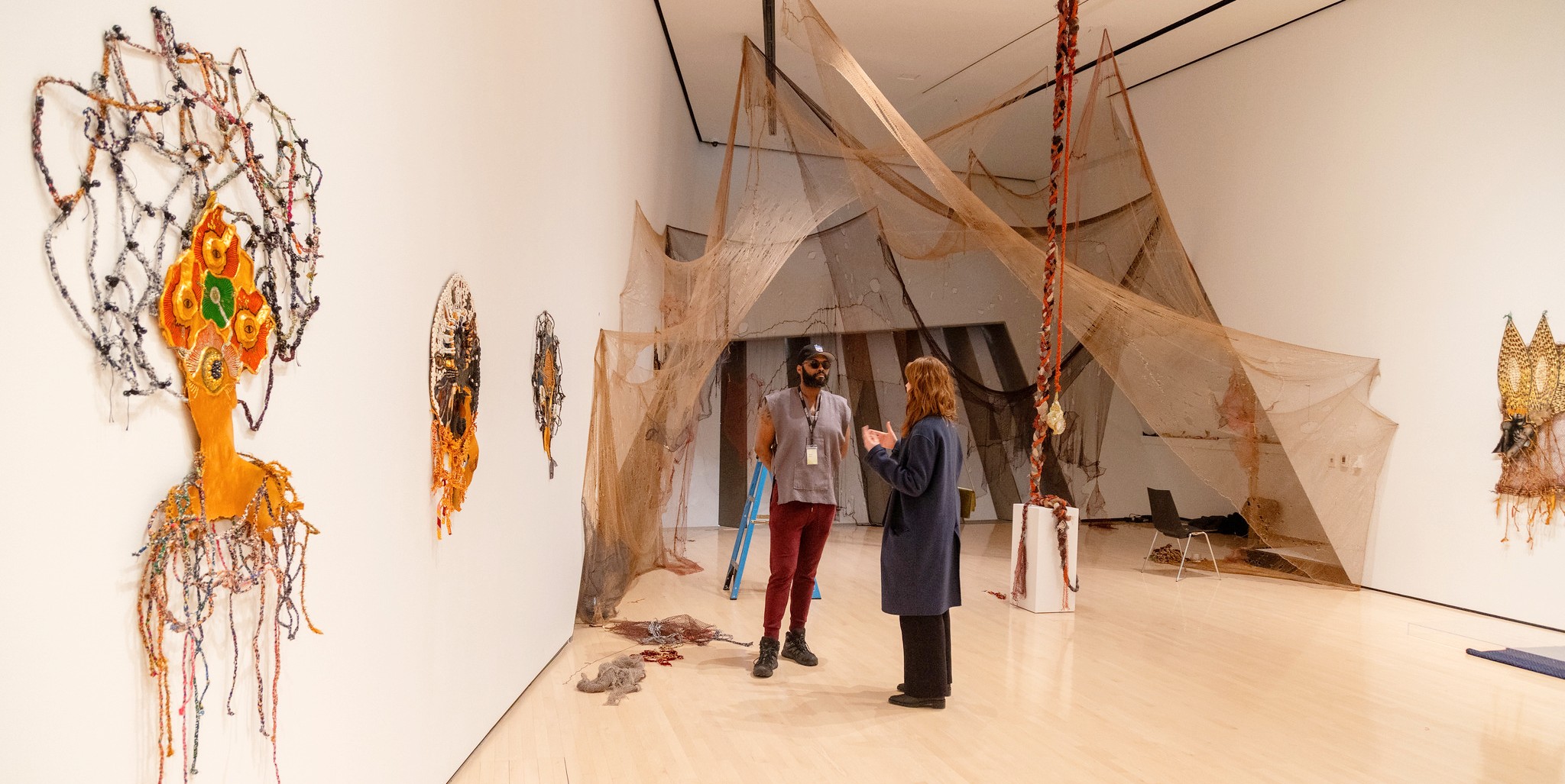 Two people engage in conversation in an art gallery. Intricate textile artworks and large, draped netting installations surround them, creating an immersive, creative atmosphere.