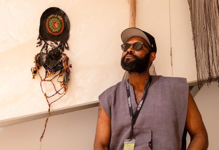 A man with a beard, sunglasses, and cap stands pensively beside an abstract wall art piece with beads. The scene exudes a creative and thoughtful atmosphere.