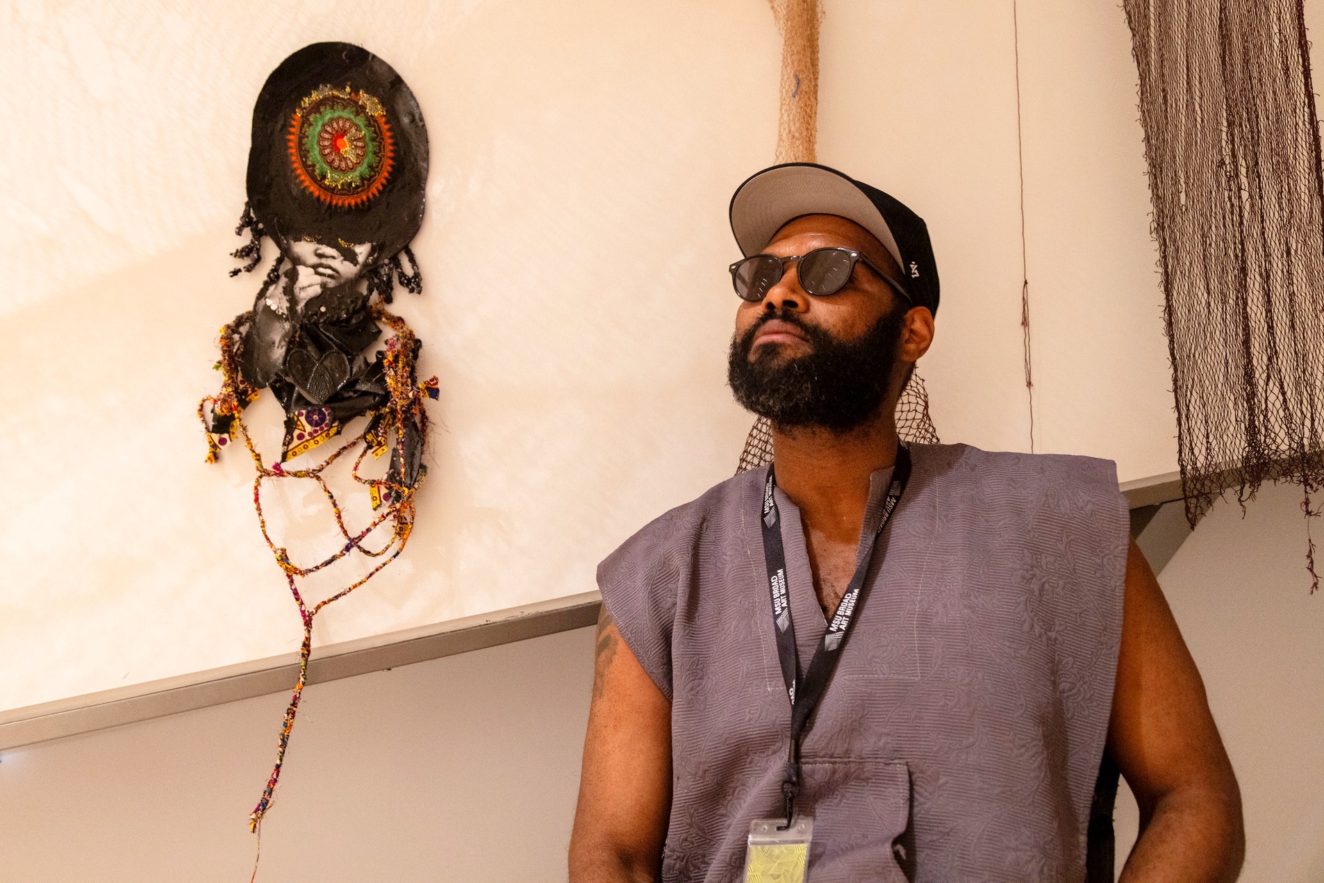 A man with a beard, sunglasses, and cap stands pensively beside an abstract wall art piece with beads. The scene exudes a creative and thoughtful atmosphere.