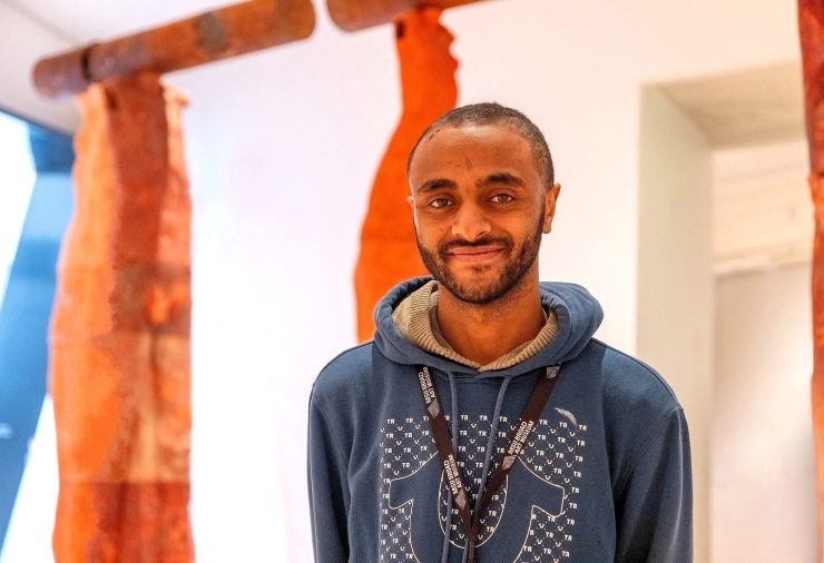 A smiling person in a blue hoodie stands in front of abstract orange and brown sculptures in a well-lit gallery space.