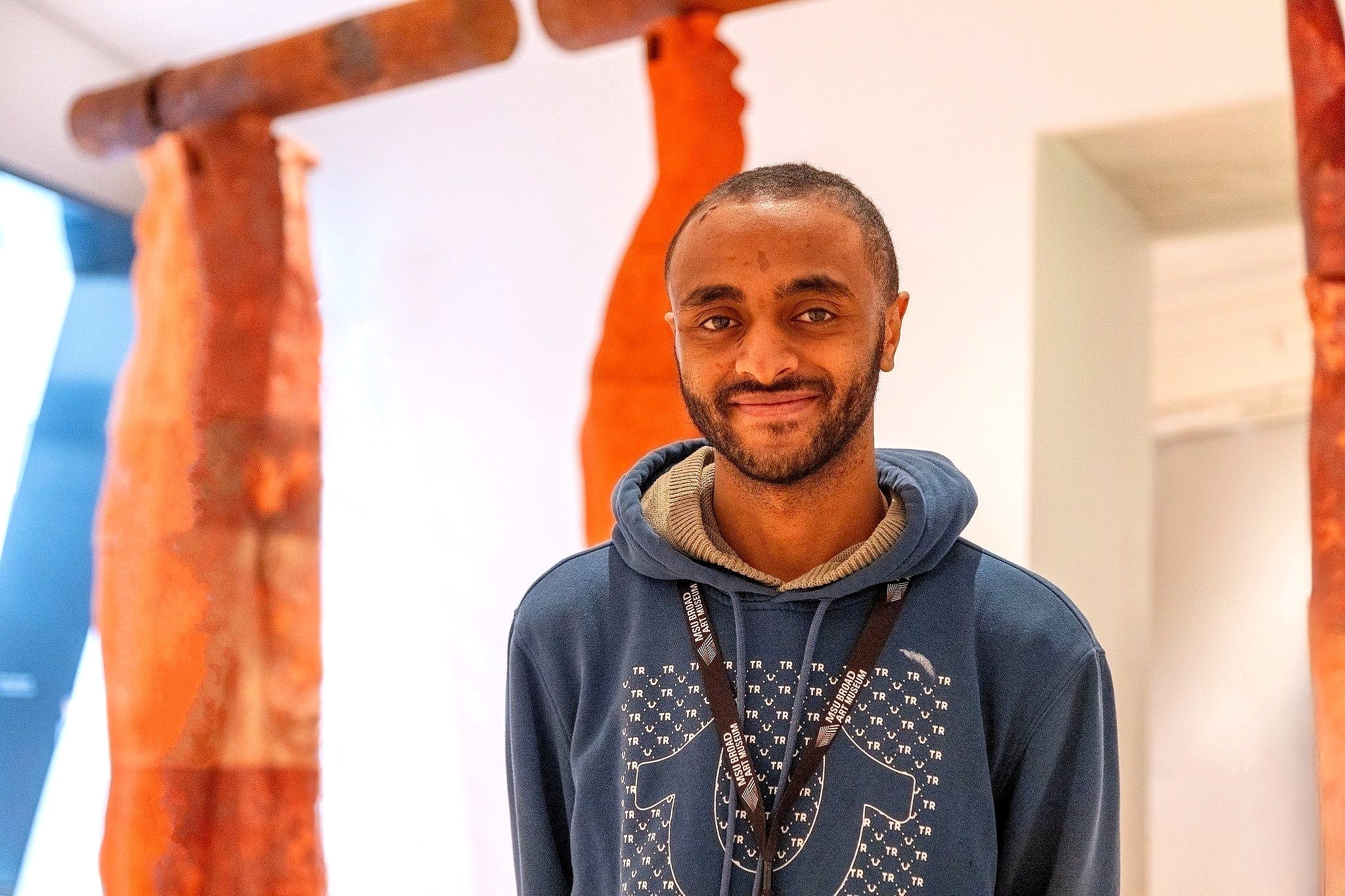 A smiling person in a blue hoodie stands in front of abstract orange and brown sculptures in a well-lit gallery space.