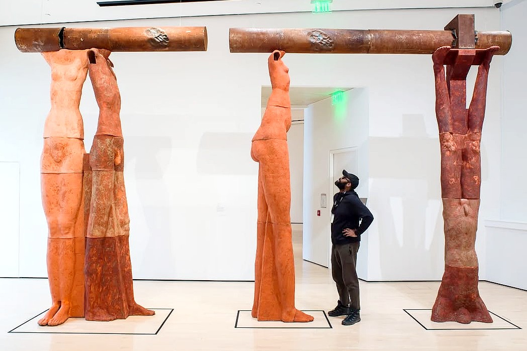 A man observes three clay human figures in a gallery. Each figure holds a textured beam overhead, creating a sense of strength and curiosity.