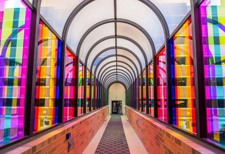 An arched walkway with glass windows covered in multicolored strips of vinyl. Through the colorful transparent windows, a parking garage can be seen.