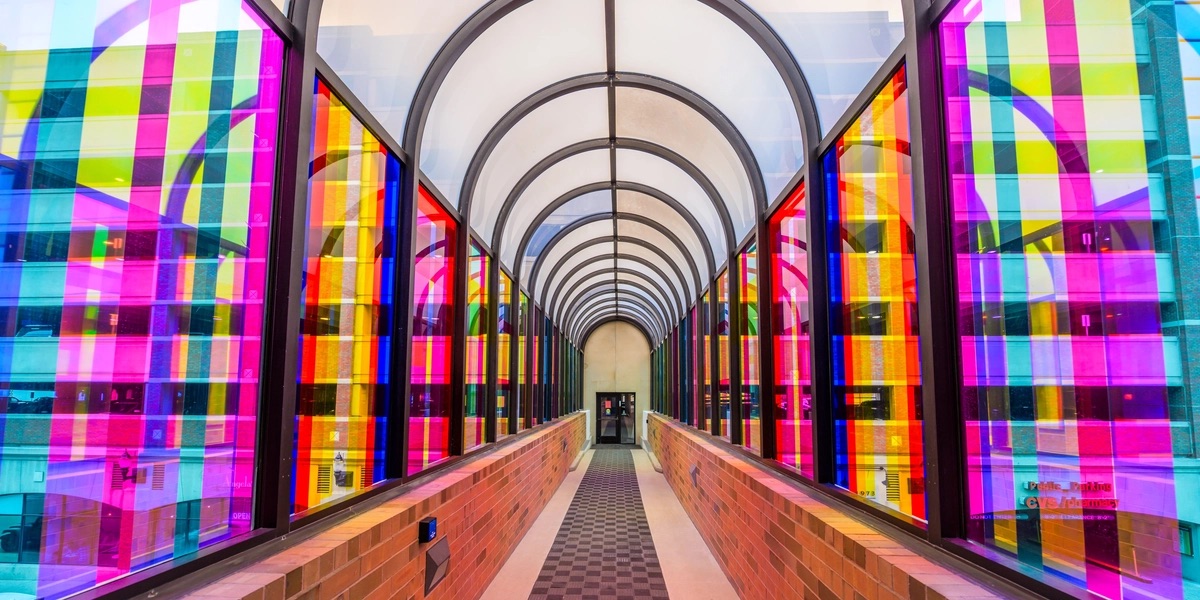 An arched walkway with glass windows covered in multicolored strips of vinyl. Through the colorful transparent windows, a parking garage can be seen.