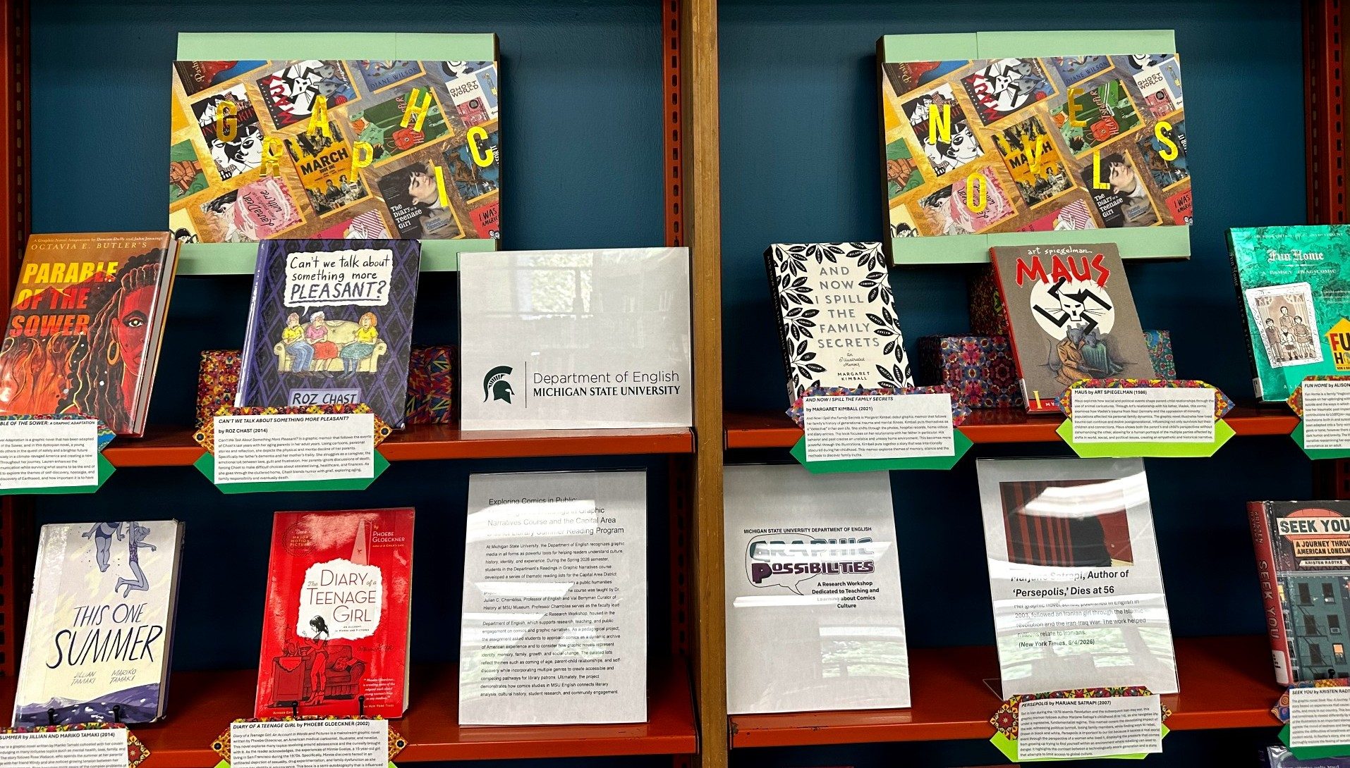 A display of graphic novels and comics-themed books arranged on orange shelving highlights a collaborative reading exhibit by Michigan State University's Department of English. Featured titles include Parable of the Sower, Can't We Talk About Something More Pleasant?, Maus, This One Summer, Diary of a Teenage Girl, and Seek You, accompanied by descriptive place cards and informal signage about graphic narratives and community reading. Decorative boards spelling 
