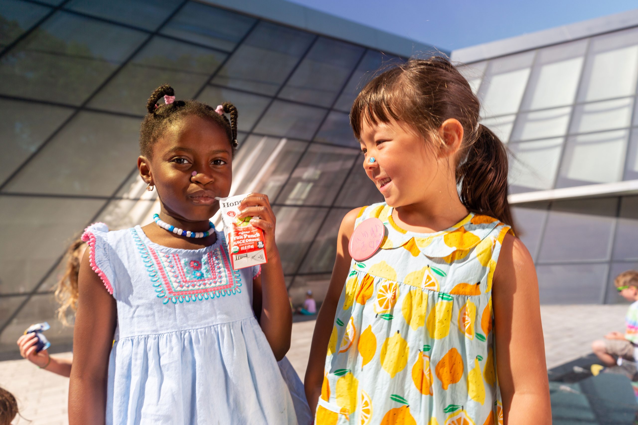A young girl with braids sips a juice box while a young girl in a yellow dress looks at her and smiles.