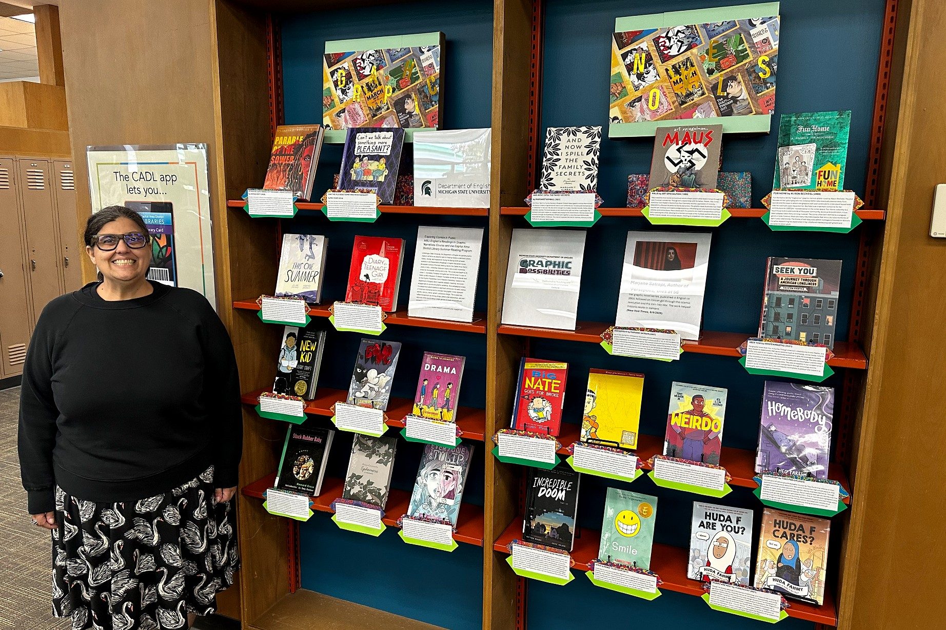 A smiling person stands beside a library display of colorful graphic novels arranged on shelves. Each book has a green label.
