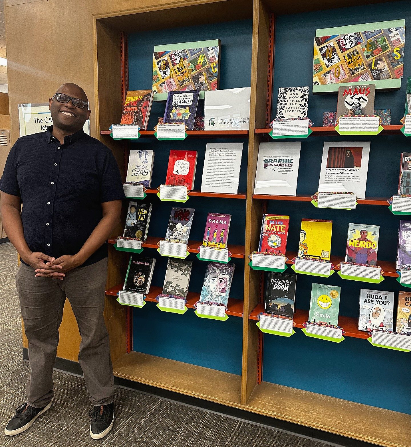 A person smiling stands beside a library display featuring various graphic novels. The shelves are filled with colorful covers, creating a vibrant and welcoming atmosphere.