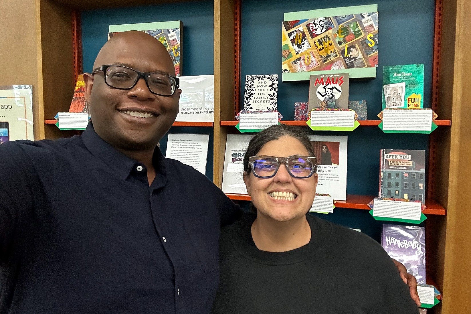 Two smiling people stand arm in arm in front of a library-style display featuring graphic novels and informational cards. Behind them, books including Maus, Fun Home, Seek You and Homebody are arranged on shelves as part of a comics-themed exhibit.