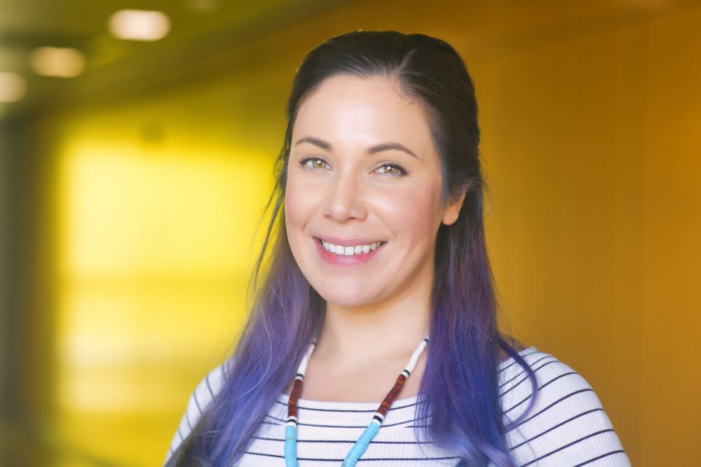 A woman with long dark hair that's dyed purple at the ends wearing a striped shirt and beaded necklace. The background is a wooded panel hallway.