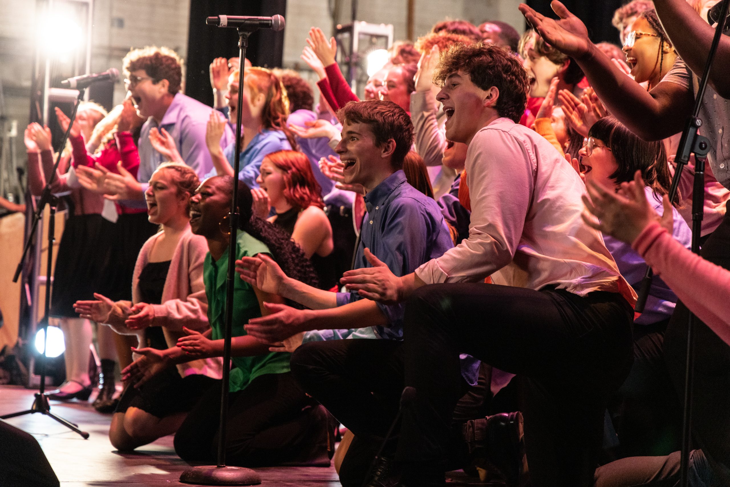Young performers group together with hands outstretched, singing on stage under lights