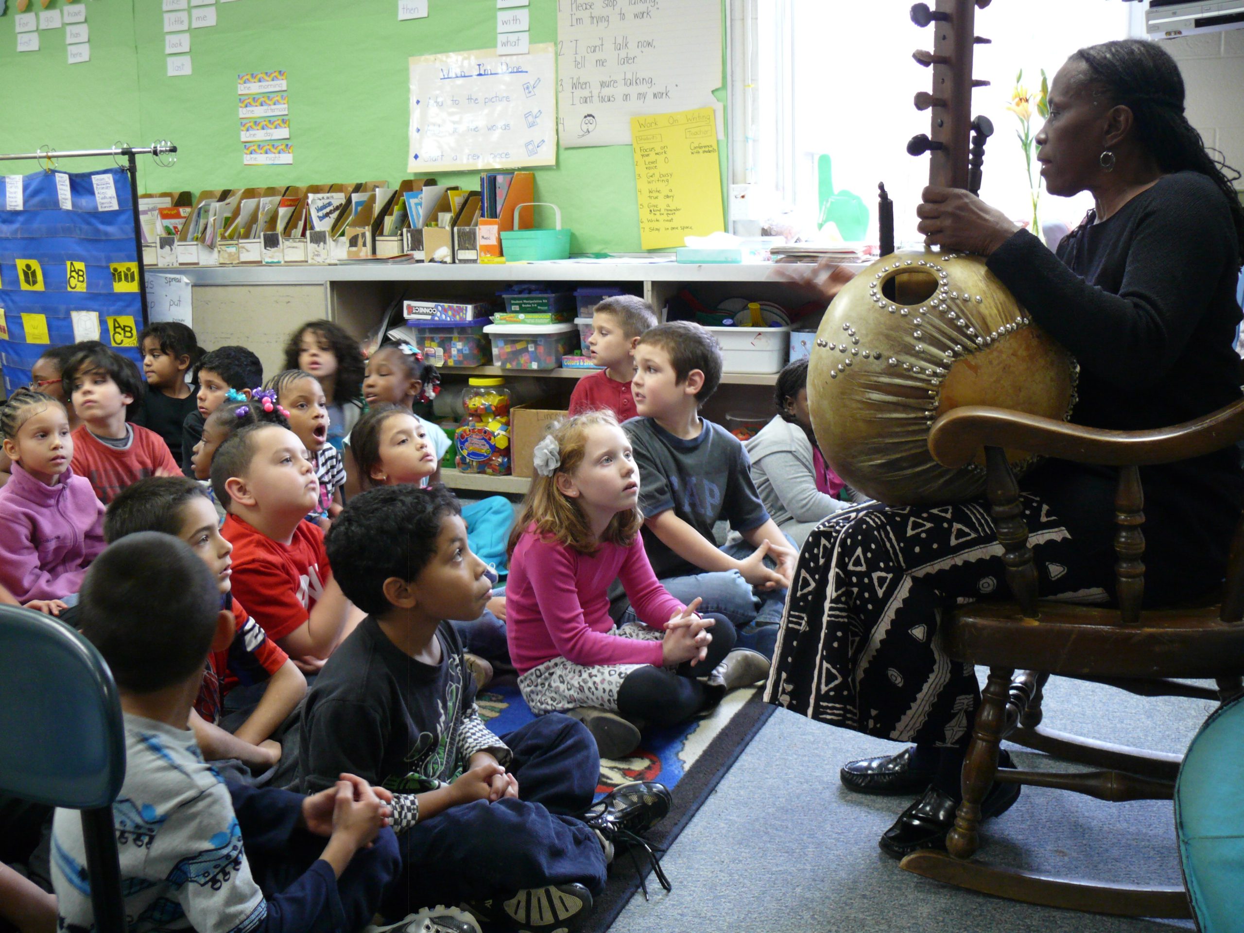 About 20 children sit in front of a woman seated in a chair showing a traditional African instrument