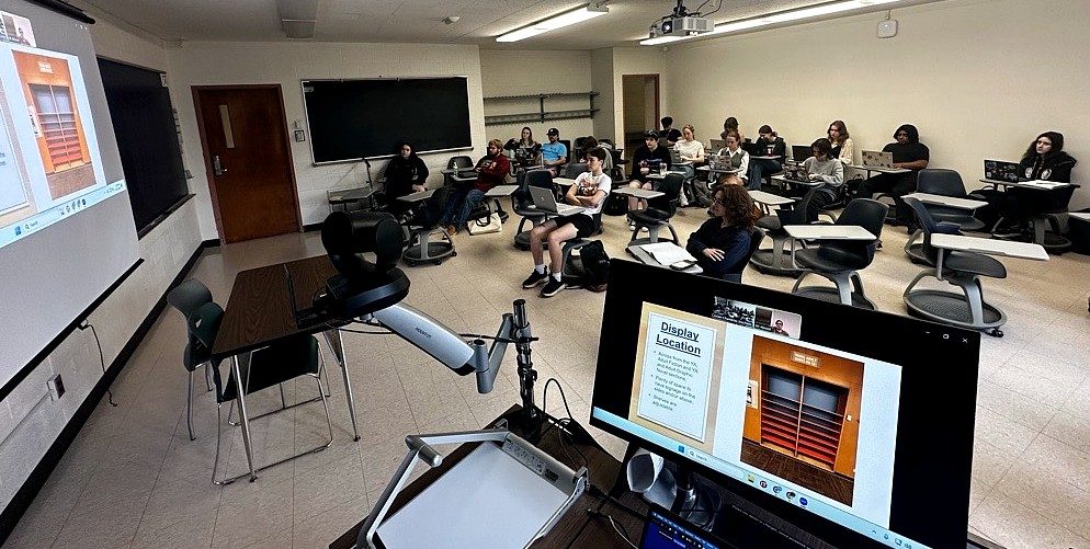 Classroom with students seated at desks, facing a large screen displaying a presentation. A camera and computer screen are in the foreground.