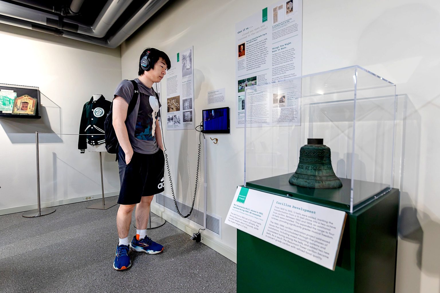 A visitor wearing headphones listens to an interactive audio display while viewing artifacts and interpretive panels in a museum exhibition on Michigan State University Traditions. A historic carillon bell is displayed in the foreground.