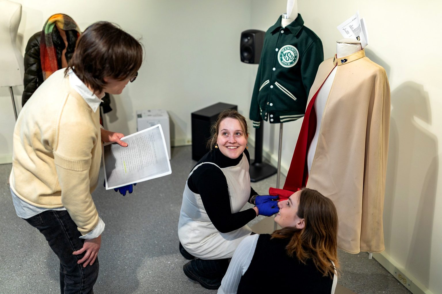 An instructor consults with two students as they install garments and interpretive materials for a museum exhibition featuring Michigan State University memorabilia, including a varsity jacket and a ceremonial cape displayed on mannequins.