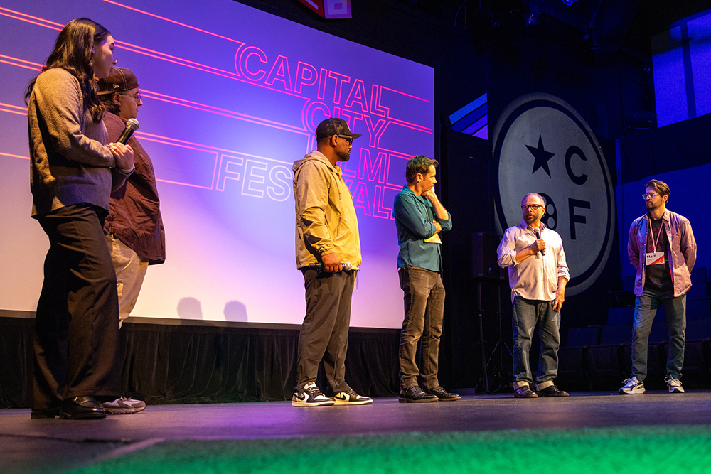 Six people stand on stage talking with a crowd about films and filmmaking. In the background are the words 