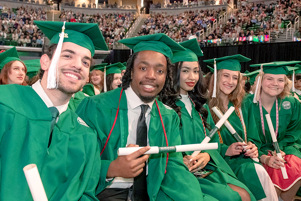 A group of five Michigan State University students wearing green caps and gowns smile while holding rolled paper diplomas in a graduation ceremony.