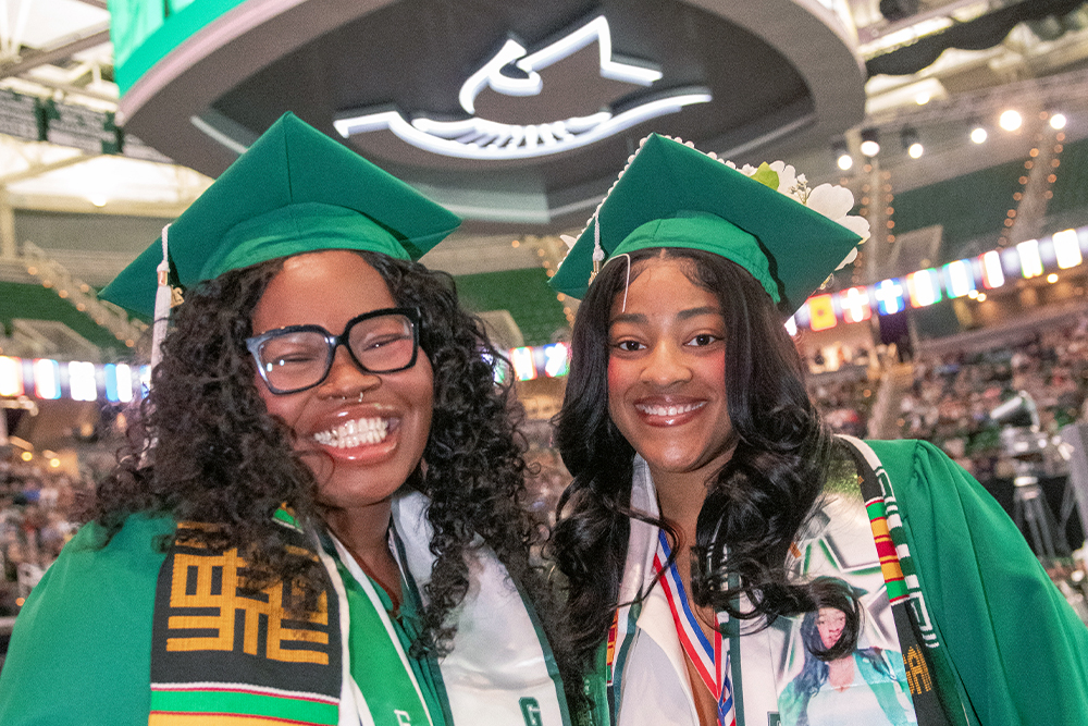 Two Michigan Sate University students wearing green caps and gowns along with colorful stoles smile in a graduation ceremony setting. Above them is a Jumbotron showing the graduation ceremony going on.