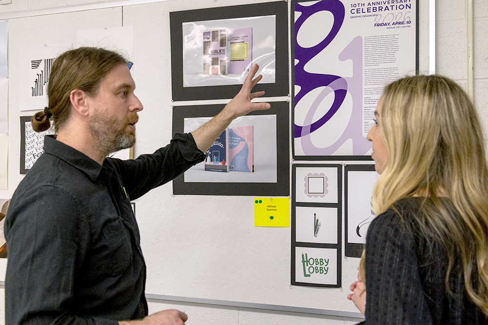A faculty member and a student, both wearing black shirts, discuss graphic design artwork displayed on a wall.