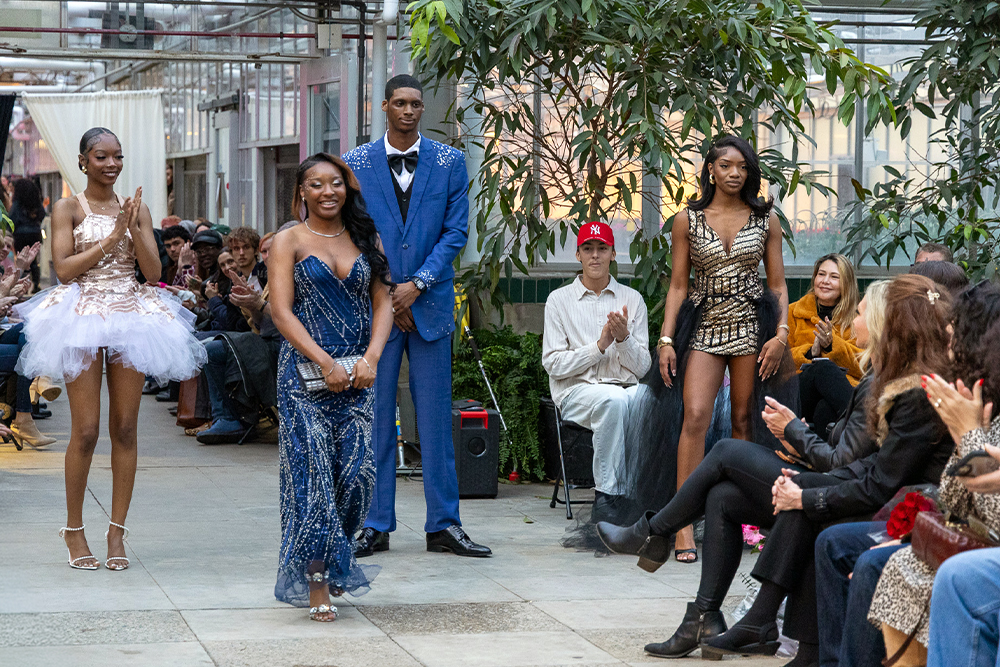 A student designer walks the runway during a fashion show in a greenhouse wearing a sparkling dark blue evening gown. Two models in formalwear, including a blue suit and a short tulle dress, stand nearby as audience members applaud.