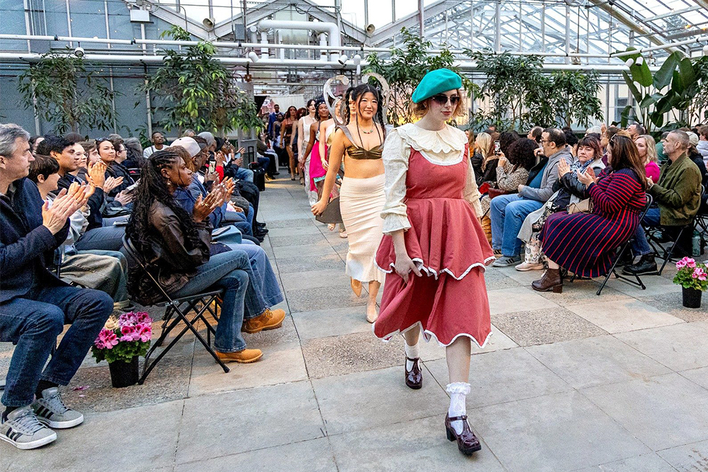 A fashion show takes place inside a large greenhouse with glass walls and ceilings. A model wearing a pink dress, green beret, and sunglasses leads a line of models down the runway as attendees sit on both sides.