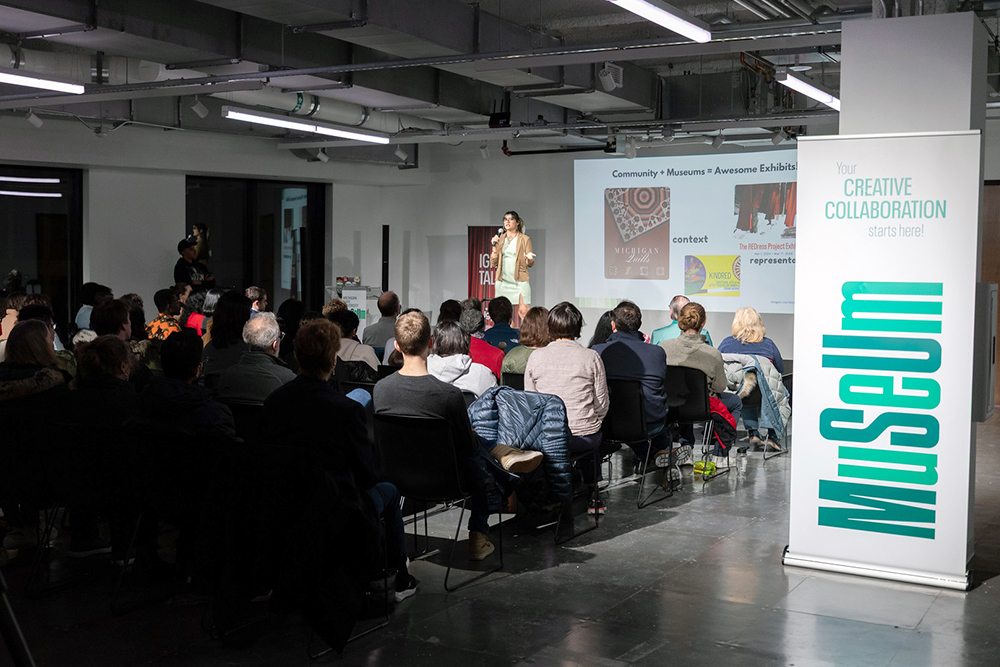 A student wearing a dress and a brown blazer stands on a stage and talks into a microphone to an audience. On the screen behind the student are examples of museum displays including Michigan Indigenous quillwork. Words on the screen say: 