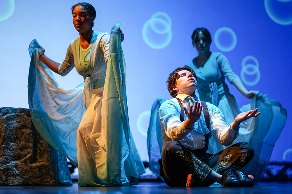 A performer sits cross-legged at center stage with palms raised upward, illuminated by cool blue lighting and circular projections. Two dancers stand behind holding flowing blue fabric.