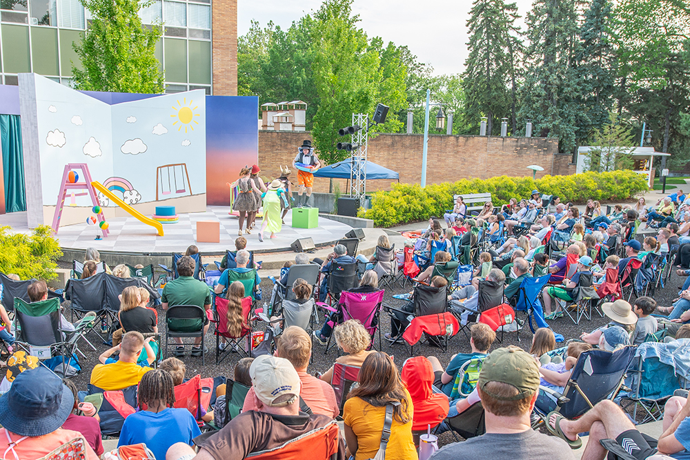 Audience members sitting in lawn chairs watch an outdoor theatre performance at Michigan State University's Summer Circle Theatre. Onstage actors perform in a scene featuring a colorful bedroom setting.