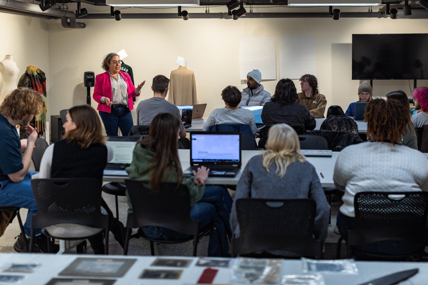 An instructor speaks to students during a museum studies class while archival materials and exhibition objects are displayed in the foreground and museum artifacts, including historical garments, are visible behind the class.