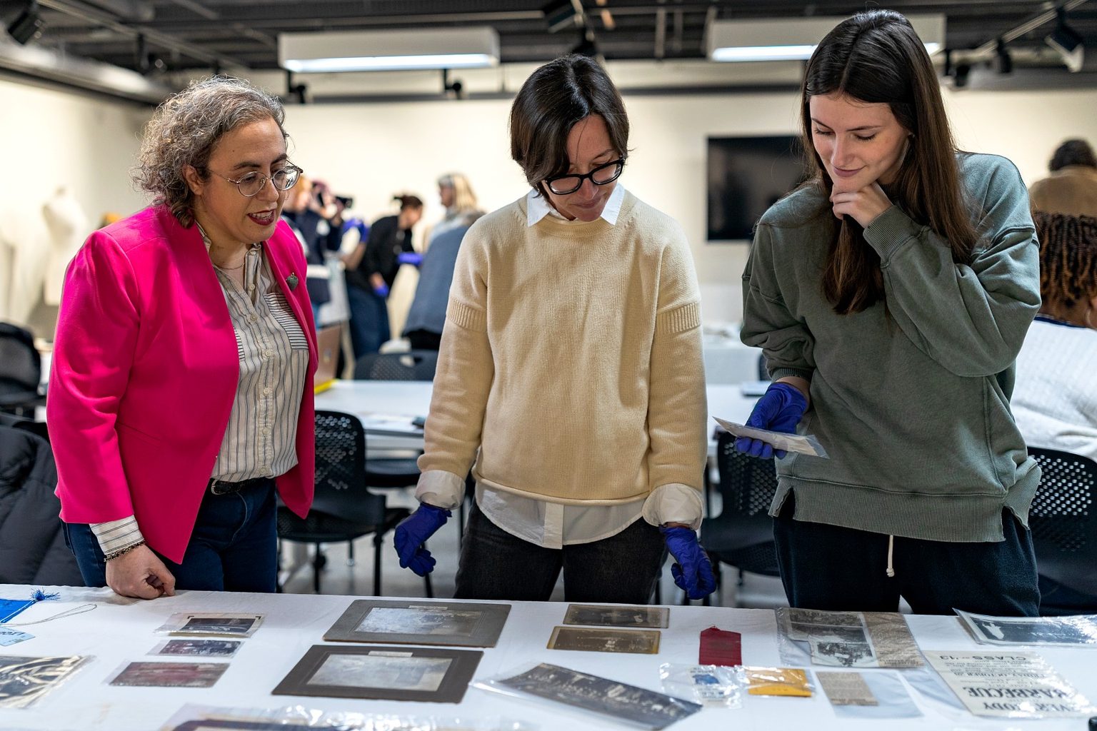 An instructor and two students wearing blue archival gloves examine historic photographs, newspaper clippings, and other artifacts spread across a table during a hands-on museum studies class.