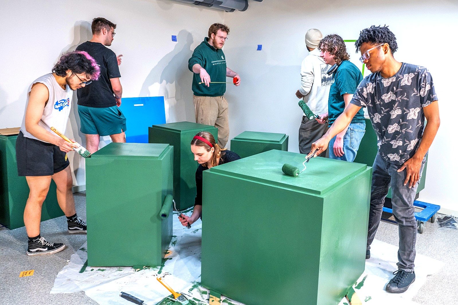 Students paint green exhibition pedestals as part of the installation process for the museum exhibition, working together to prepare display furniture for the gallery.