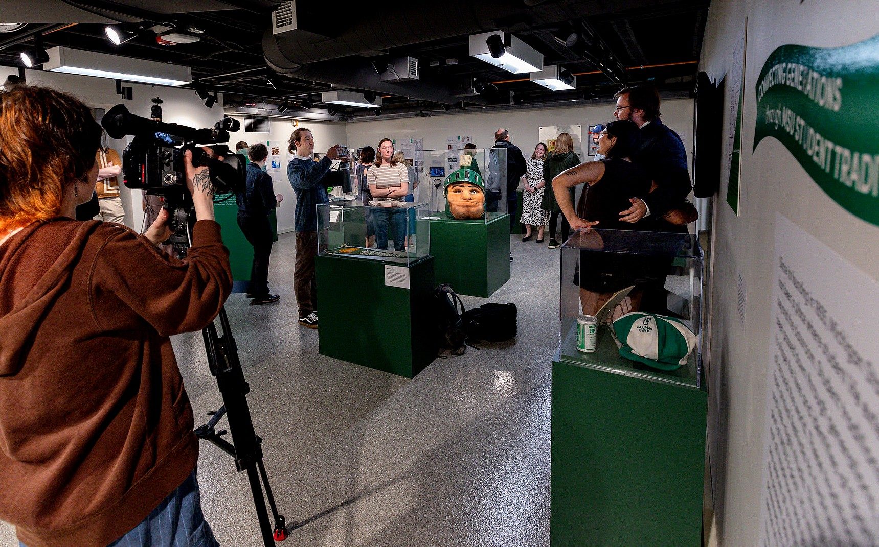 A television news crew films visitors exploring the student-curated museum exhibition on Michigan State University traditions, with artifacts, display cases, and interpretive panels throughout the gallery.