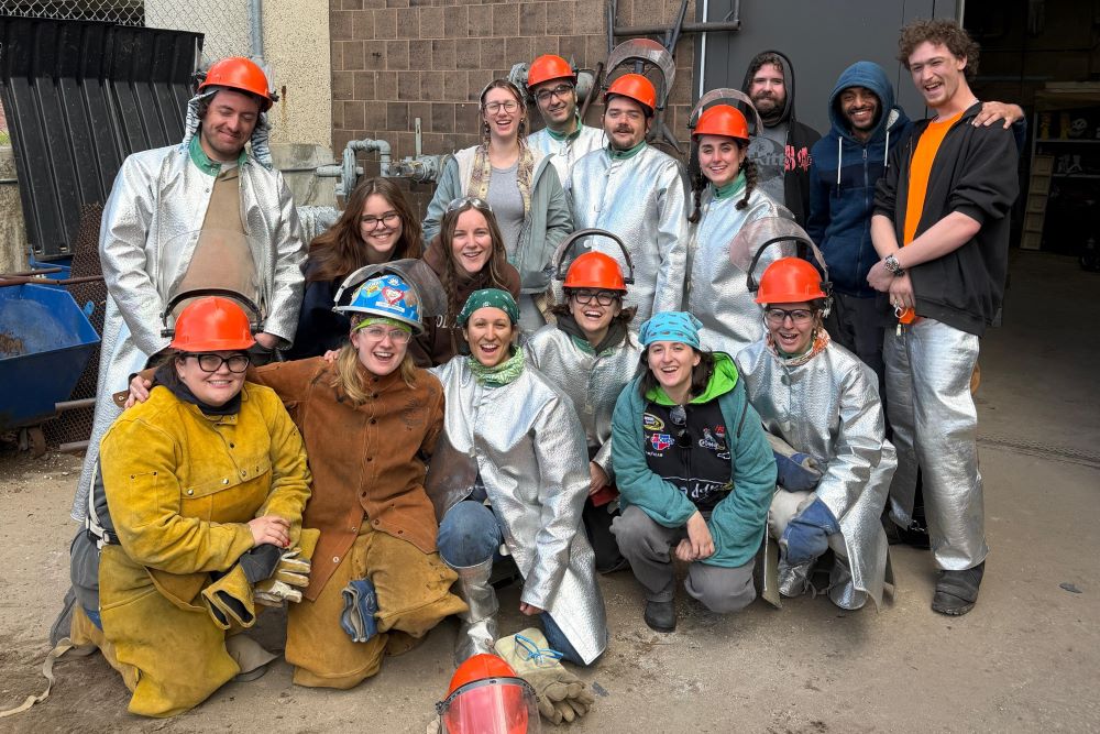 A group of 16 people take a photo together outside a foundry setting. Many of the people are wearing helmets and leather or Kevlar safety jackets used in metal casting.