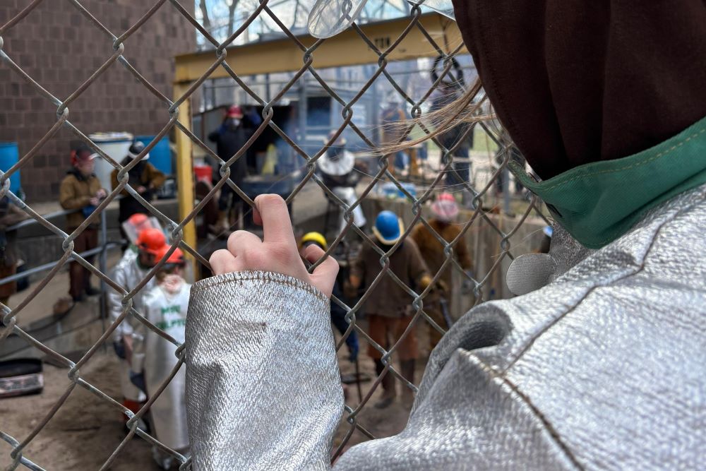 A person in a Kevlar jacket looks through a chain-length fence at people participating in an iron pour.