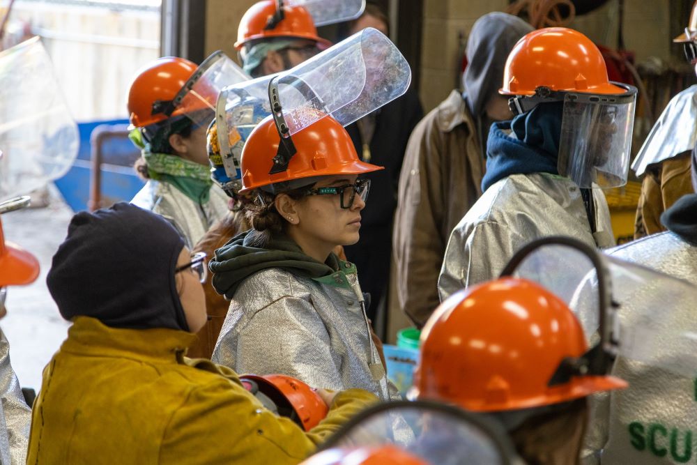People in a foundry setting wear helmets and safety jackets and listen to instructions before an iron pour event.