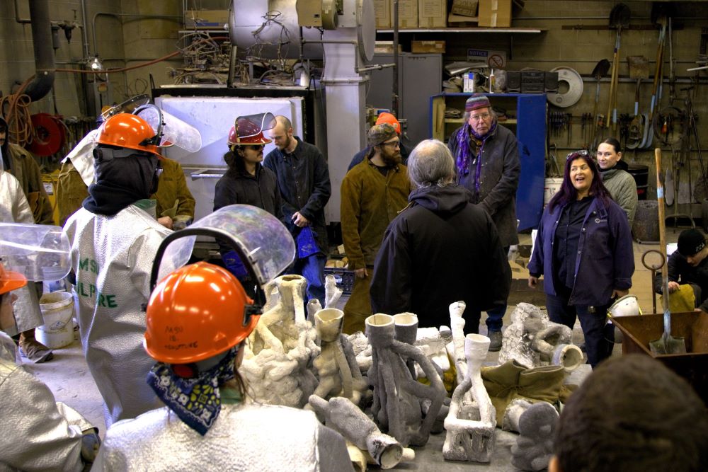 A group of people in a foundry setting, many of helmet and safety jackets, listen to instructions before an iron pour.