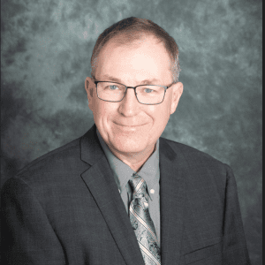 Ken Foster wearing a dark grey suit and glasses with a grey backdrop.